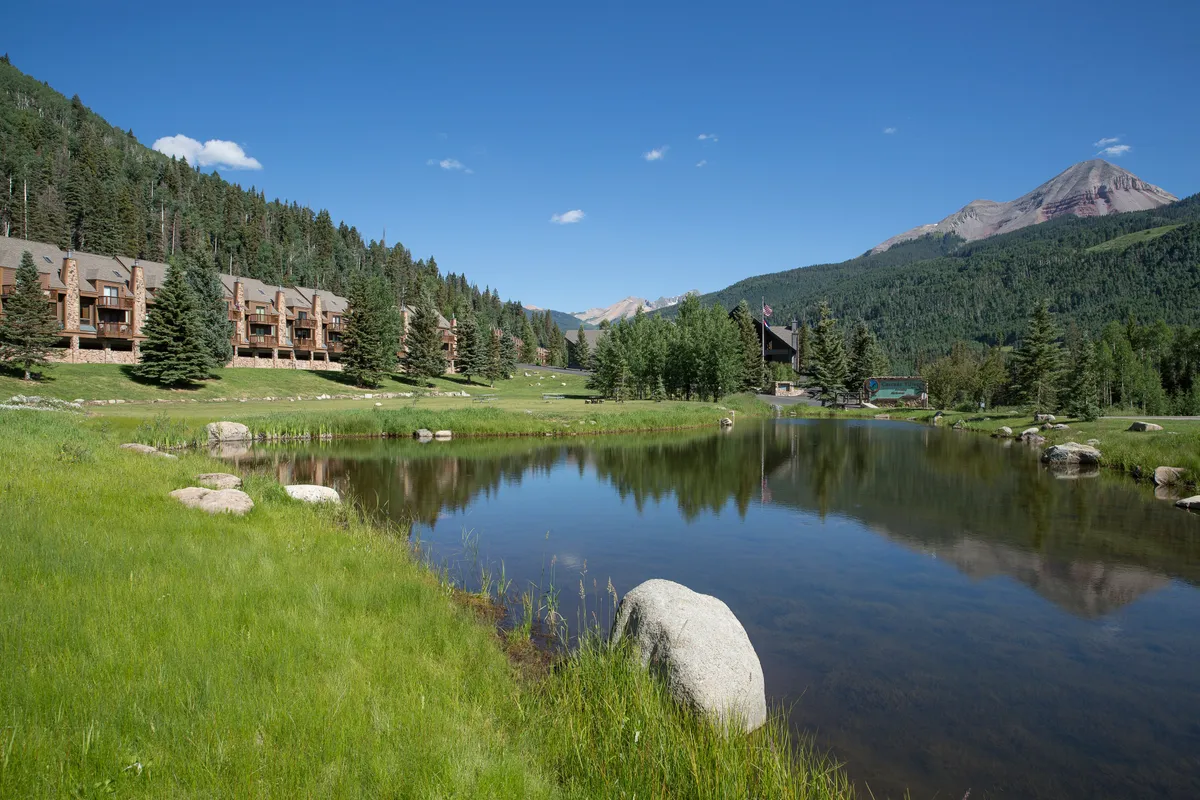 An exterior perspective featuring lodge-style buildings nestled beside calm water, with the dramatic backdrop of mountains and a lush treeline. The water mirrors the landscape, adding to the serene winter retreat ambiance.