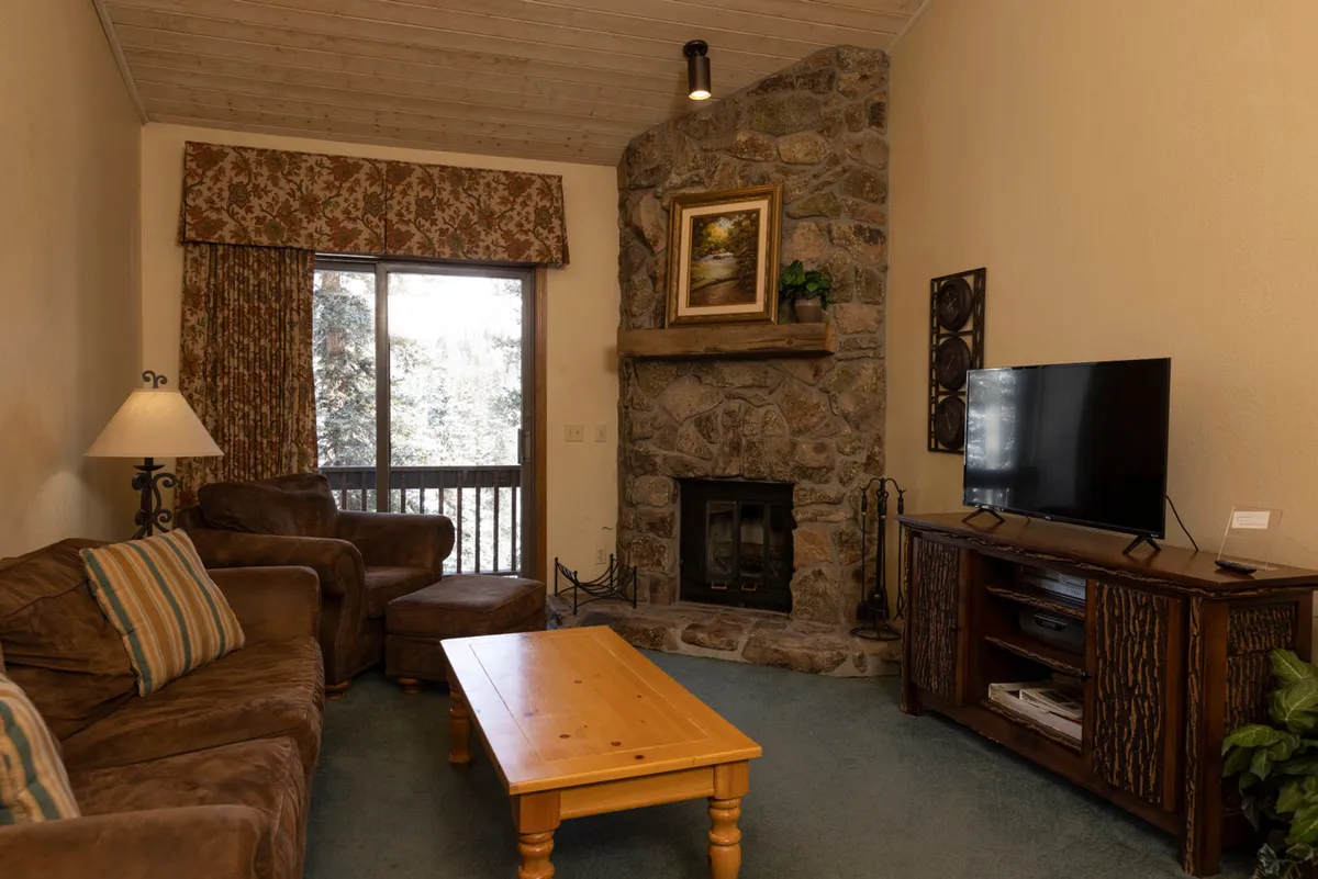 A comfortable living area centered around a fireplace, with a plush sofa, coffee table, and television. The seating is arranged to facilitate group conversation or movie nights, and the ambiance is both cozy and inviting. Large windows suggest the possibility of mountain views, enhancing the sense of place.