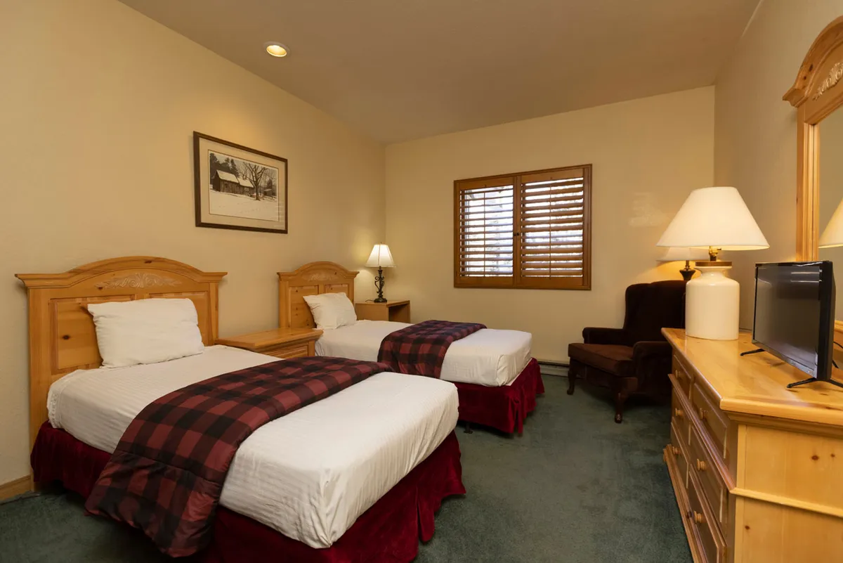A cozy second bedroom with two twin beds dressed in plaid blankets and wooden furniture, complemented by soft lighting and a window with shutters. The room’s arrangement encourages relaxation, reading, or unwinding with friends or family.