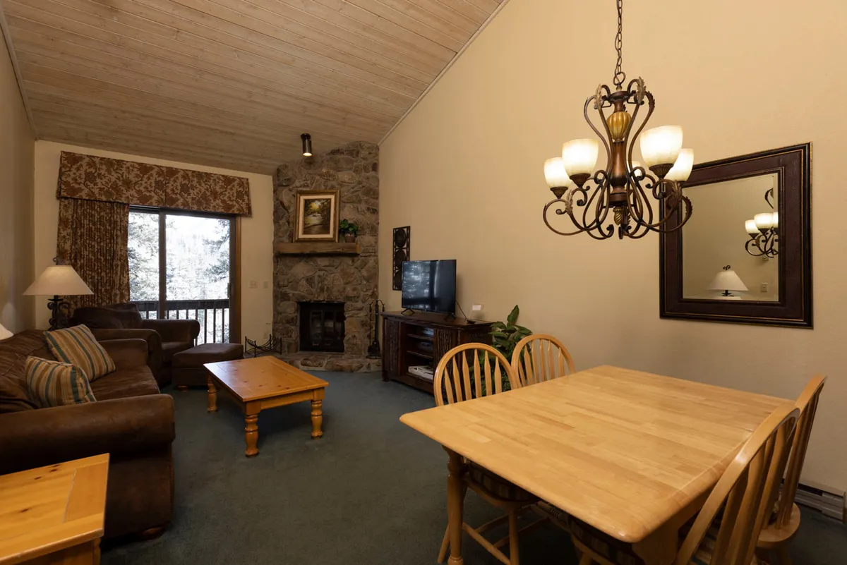 A spacious open-concept living and dining area featuring a cozy fireplace, an inviting sofa, a family-style dining table, and a warm chandelier overhead. Large windows line the wall, likely framing scenic mountain views just beyond, and the arrangement encourages socializing, relaxation, and group gatherings.
