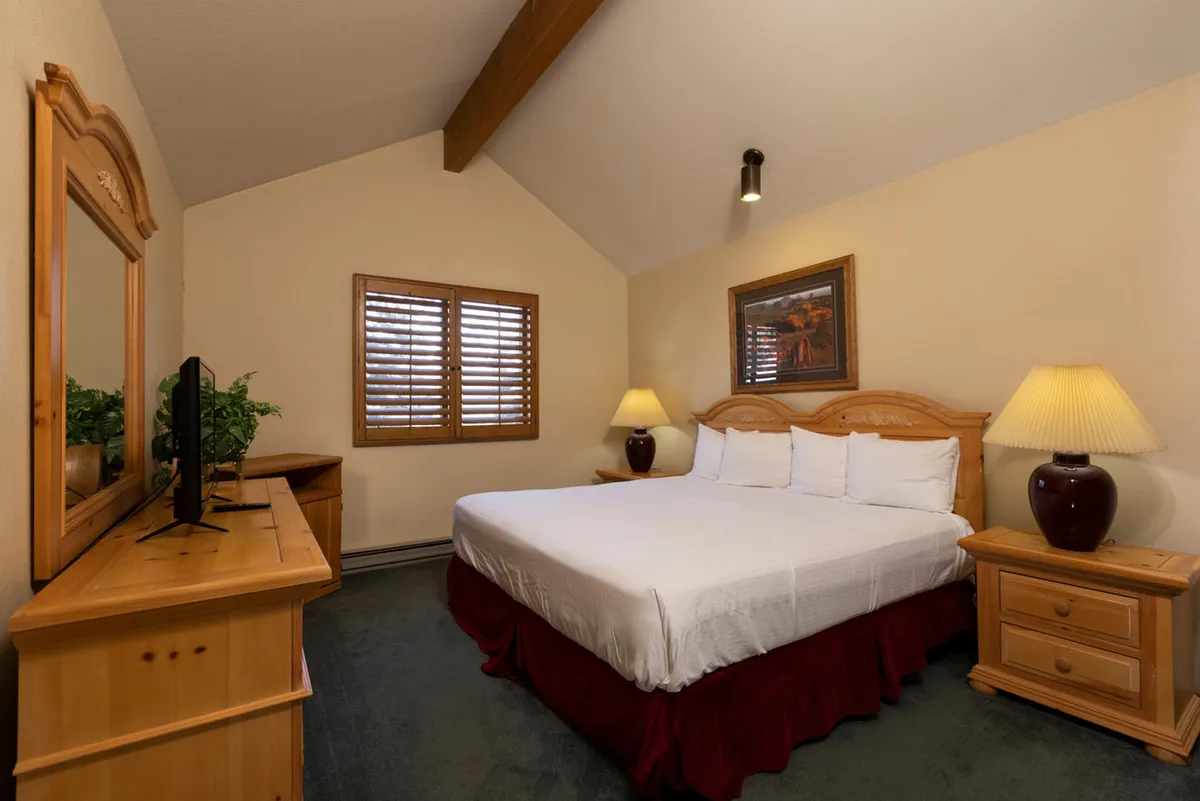 A welcoming primary bedroom featuring a comfortable bed with plush bedding, flanked by nightstands and table lamps. The space includes a dressing table and a window with shutters, creating a tranquil spot for reading or simply enjoying a peaceful atmosphere.