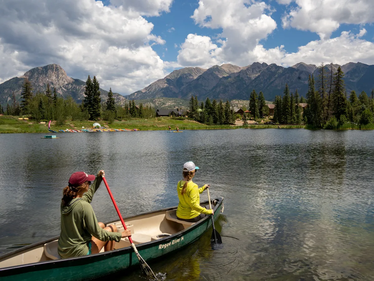 An inviting lakeside scene at sunset, featuring a group enjoying a canoe ride on a tranquil mountain lake surrounded by trees. In the background, rugged mountains rise up, and the warm glow of the setting sun creates a sense of adventure and relaxation. People are actively engaging in canoeing, while others might be fishing or picnicking nearby, all immersed in the natural beauty.