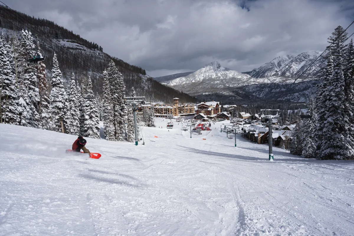 A skier in action on a snowy slope, surrounded by snow-covered trees and a majestic mountain backdrop. The ski resort buildings are visible at the base, indicating a lively and adventurous atmosphere ideal for family fun and mountain exploration.