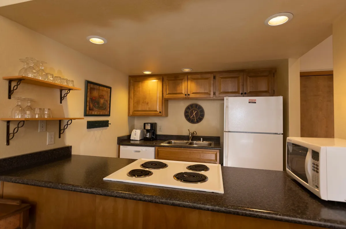 A warm, inviting kitchen showcases a spacious island at the center, surrounded by wooden cabinets, an oven and stove, and a charming wall clock. The island is uncluttered and ready for meal prep or casual gatherings, while the entire space glows with natural and artificial light, evoking a sense of welcoming coziness.