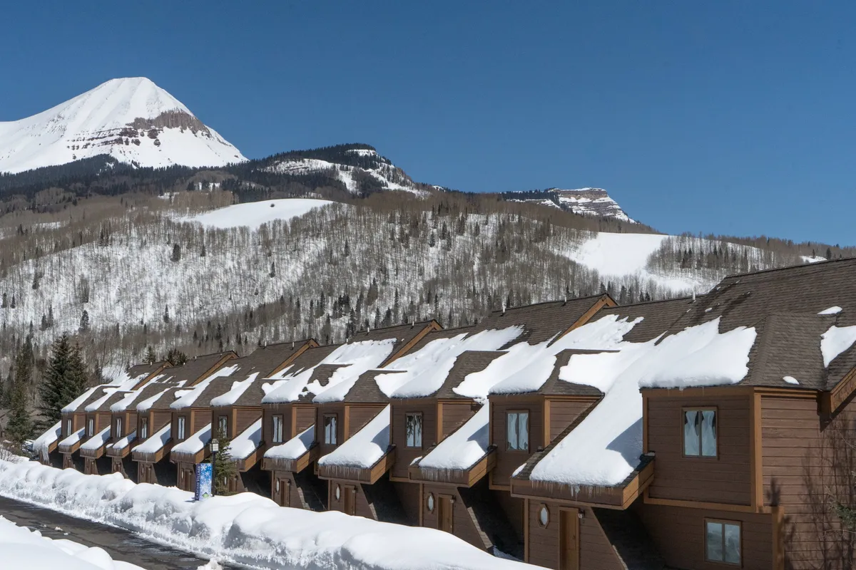Snow-covered rooftops of mountain lodges with a stunning mountain backdrop under a clear blue sky. The scene exudes tranquility and hints at cozy après-ski gatherings after days filled with outdoor activities.