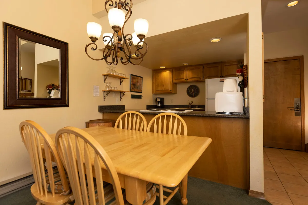 A warmly lit dining area with a wooden table, elegant chandelier, and kitchen in the background. The setup is ideal for shared meals, celebrations, or a quiet coffee morning, with a seamless flow to the adjacent living space visible in the composition.