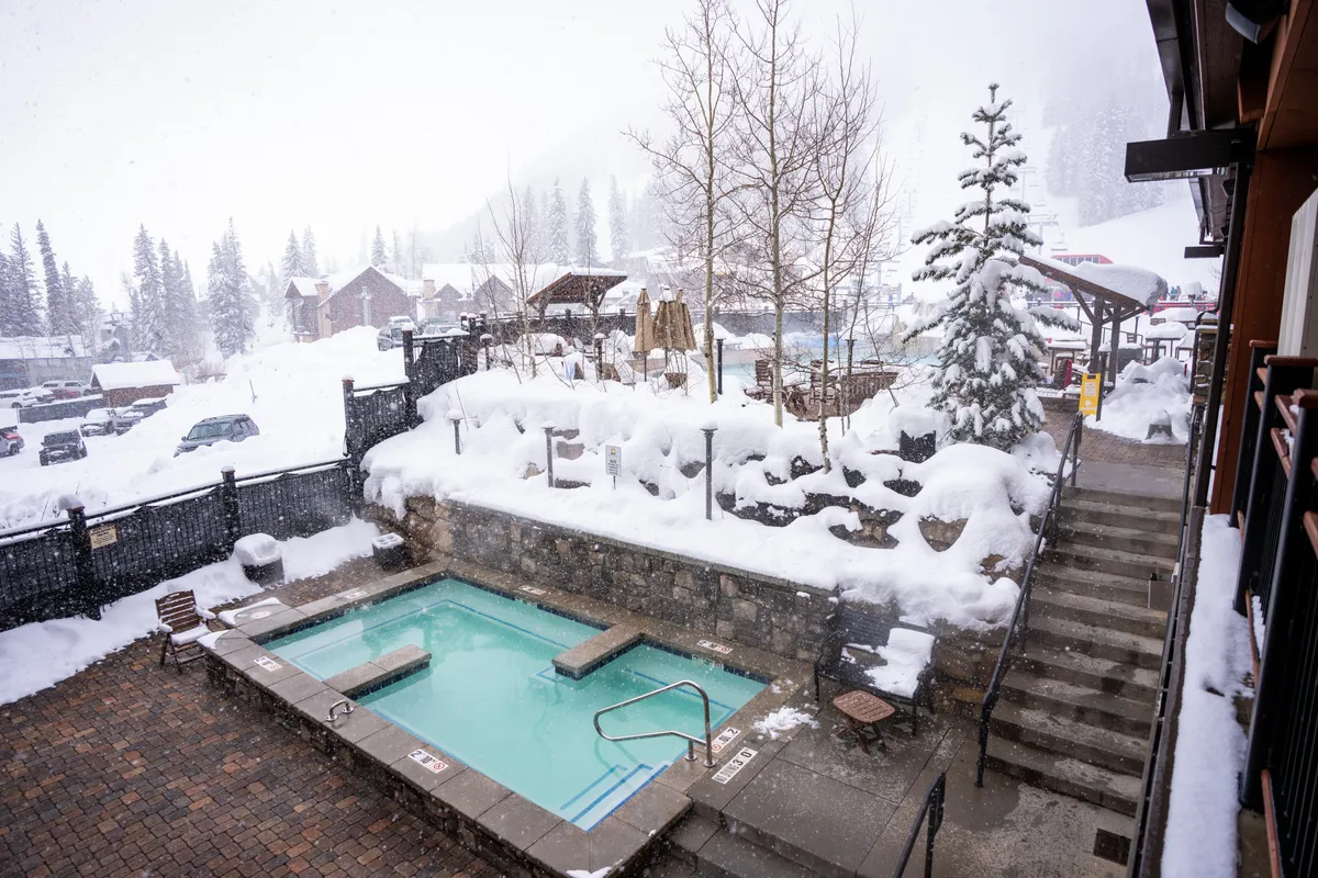 This exterior image features a steaming hot tub set amidst a snow-covered landscape, with trees and a stairway nearby. The seating area creates a cozy space to unwind while enjoying panoramic winter views.