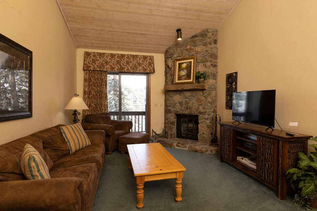 The image showcases a cozy condo living space featuring a striking stone fireplace as the centerpiece, surrounded by plush, comfortable seating. A large window lets in natural light and frames scenic mountain views, while a sturdy wooden coffee table invites guests to gather around for games, snacks, or hot cocoa. The overall ambiance is warm and inviting, perfect for relaxation and family time after a day of adventure in Durango.